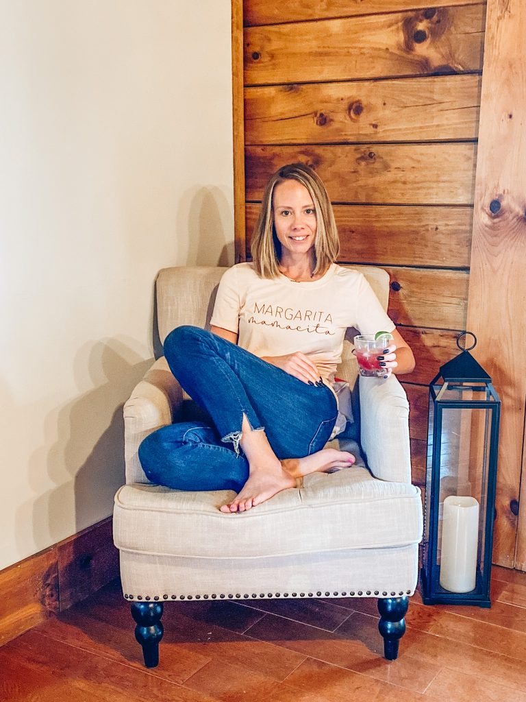 Cranberry Margarita by popular Pittsburg lifestyle blog, Fashionably Late Mom: image of a woman sitting in a grey armchair next to a tall black lantern with a pillar candle inside and holding a cranberry margarita. 