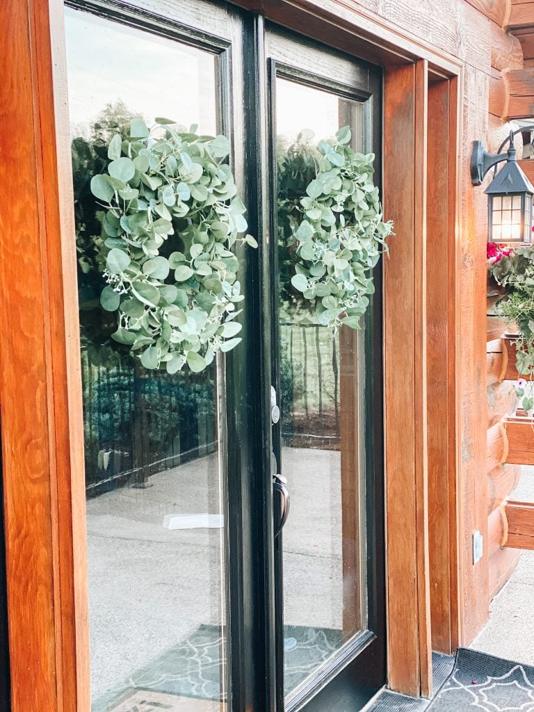 eucalyptus wreaths |  Summer Front Porch by popular Pittsburg life and style blog, Fashionably Late Mom: after image of a front porch on a log home with black frame doors, black planters with pink flowers, hanging potted pink flower planters, and greenery wreaths. 