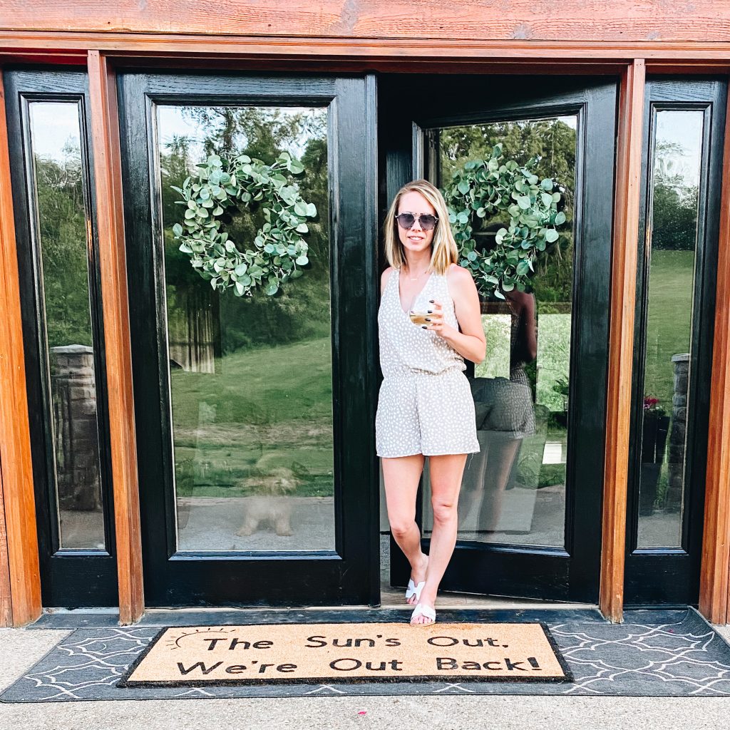 front porch decor, doormat |  Summer Front Porch by popular Pittsburg life and style blog, Fashionably Late Mom: image of a woman holding a glass of wine and standing on her front porch on a log home with black frame doors, black planters with pink flowers, hanging potted pink flower planters, and greenery wreaths. 