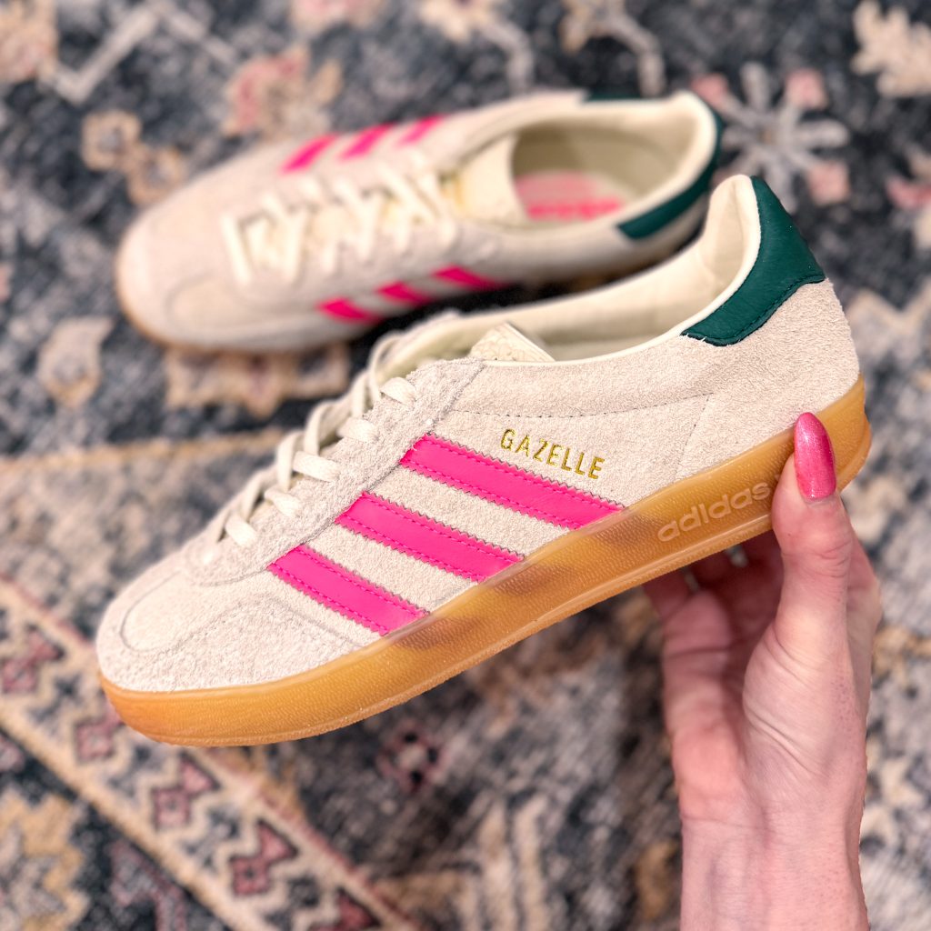 A close-up image of a pair of Adidas Gazelle sneakers featuring a cream upper with pink stripes and a green accent on the heel, being held against a patterned rug.
