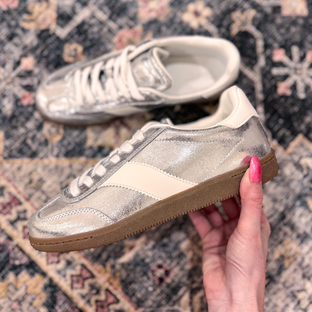 Silver statement sneakers featuring a metallic finish, paired with a subtle cream accent and gum sole, held against a patterned background.